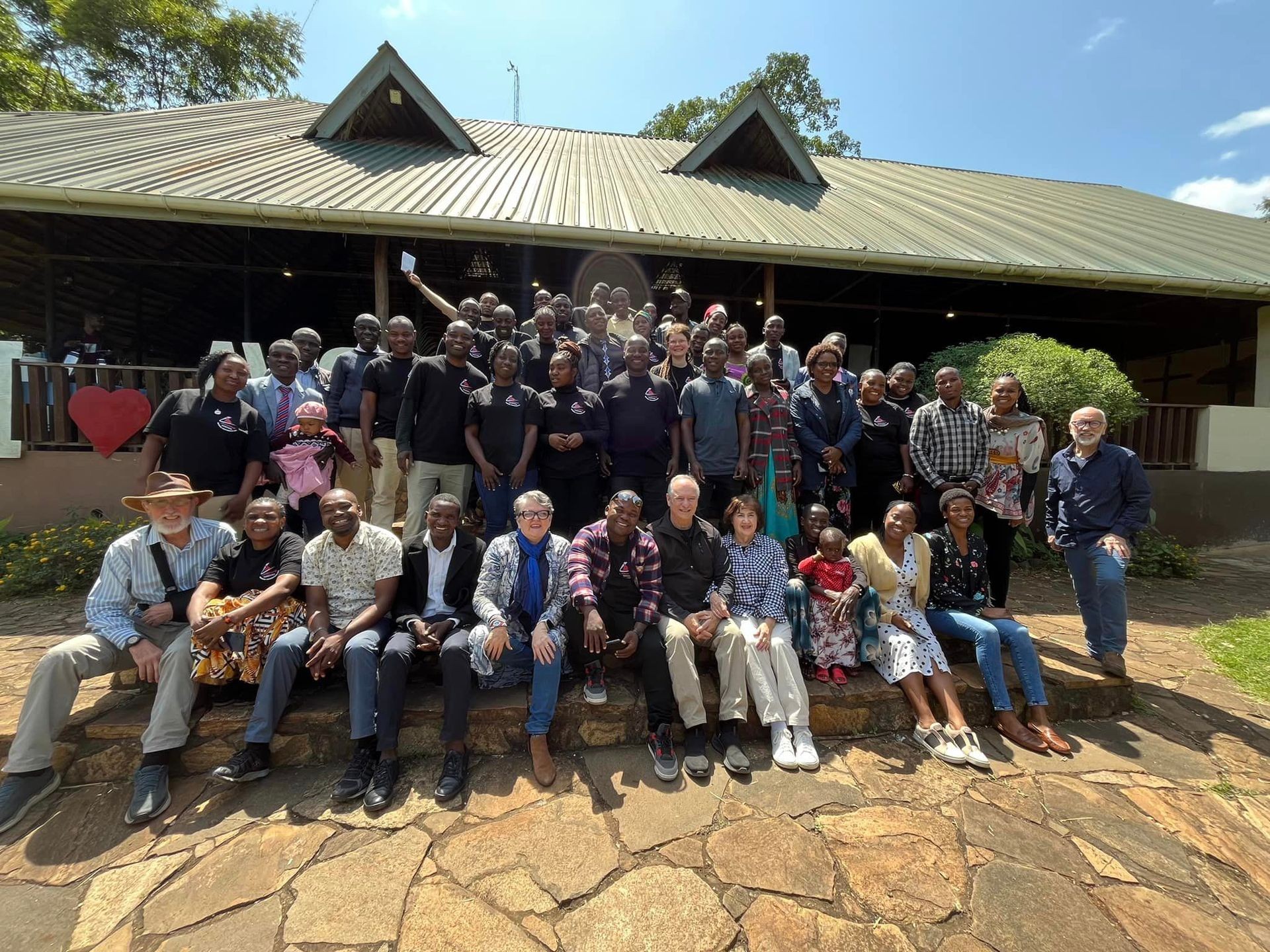 Large group of people posing in front of a building with a tin roof on a sunny day.